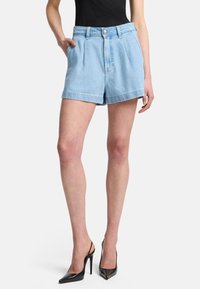 Shorts in denim blu chiaro con vita alta, caratterizzati da pieghe frontali e tasche laterali, abbinati a tacchi alti neri a punta.