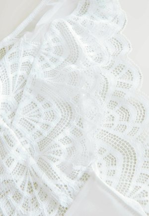 Bílá krajková látka s jemnými květinovými a geometrickými vzory, s vlnkovým okrajem na hladkém neprůhledném panelu. Lehké textury.