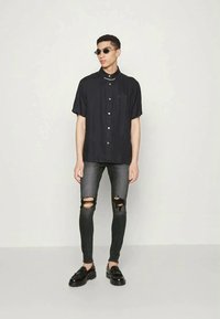 Camisa preta de manga curta com fecho de botão, combinada com jeans skinny escuros rasgados e sapatos pretos. Modelo a usar óculos de sol.