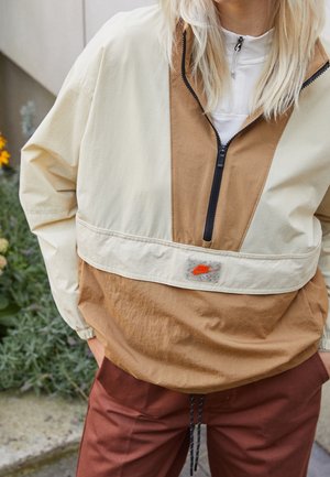 Personne portant une veste Nike beige et marron à demi-fermeture éclair, les mains dans les poches, debout en plein air près de verdure et de fleurs.