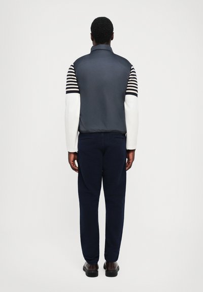 Marineblaues Weste mit gestreiften weißen Ärmeln, hohem Kragen, getragen über marineblauen Hosen und schwarzen Schuhen; glattes Material und taillierter Schnitt.