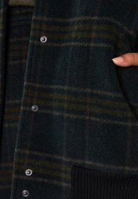 Cardigan di lana a scacchi verdi con bottoni a pressione argento, rifiniture a coste in nero sulle tasche e un tessuto morbido e strutturato.