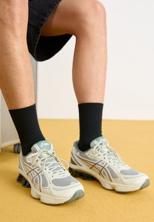 Lichtgrijze sportschoenen met mesh en synthetische overlays, voorzien van groene accenten en een gebogen zwarte gelzool, gedragen met zwarte sokken.