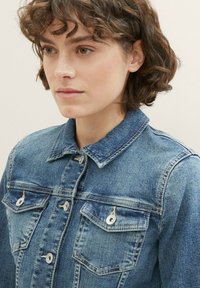 Giacca in denim di colore azzurro chiaro con colletto, chiusura frontale con bottoni e due tasche sul petto con dettagli metallici. Tessuto strutturato.