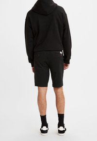 Haut à capuche noir, short noir et chaussettes noires avec des baskets blanches. Le short possède des poches latérales et arrière. Tissu lisse, design décontracté.