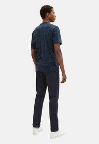 Mörkblå t-shirt med kort ärm och en strukturerad, melerad mönster, i kombination med mörka byxor och vita sneakers, sedd bakifrån.
