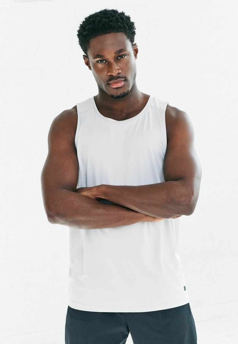 Weißes Sporttanktop mit glatter Textur, runder Ausschnitt und weiten Armausschnitten, kombiniert mit dunklen Shorts.