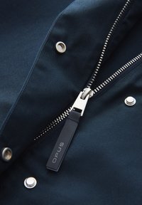 Marineblauwe jassenstof met een opvallende rits, zilverkleurige hardware, drukknoopjes en een label met de tekst "OPUS" nabij de rits.
