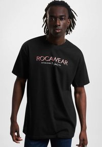 Fekete, rövid ujjú póló, amely fehér és piros "ROCAWEAR" logóval és a megalapítás dátumával készült, puha pamut anyagból.