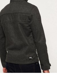 Veste en mélange de laine gris foncé avec un col montant, surface texturée et étiquette de marque près de l'ourlet, présentant une coupe ajustée et des détails de boutons.