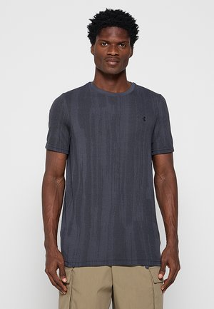 Mężczyzna w ciemnej, teksturowanej koszulce z krótkim rękawem i beżowych spodniach typu cargo, stojący na jasnym, jednolitym tle.