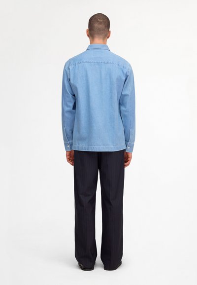 Homme debout de dos portant une chemise en denim bleu clair et un pantalon sombre à fines rayures sur un fond blanc uni.