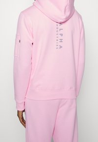 Person som bär en ljusrosa hoodie och matchande byxor, med vertikal "Alpha Industries" text på ryggen av hoodien.