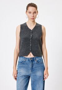 Grijze, mouwloze, gebreide top met een v-hals, voorzien van een knoopsluiting aan de voorkant en een cropped, asymmetrische zoom. Gecombineerd met lichtblauwe jeans.