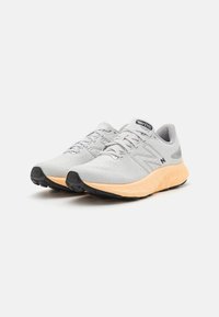 Jasnoszare buty sportowe z textured mesh górą, pomarańczową piankową środkową podeszwą i czarną gumową podeszwą. Posiadają sznurowadła i detale z logo.