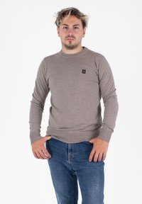Maglione a maglia marrone chiaro con maniche lunghe, collo rotondo e polsini a coste, abbinato a jeans in denim blu. Presenta un piccolo logo sul petto.