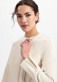 Witte leren jas met een hoge kraag, ritssluitingen aan de voorkant en mouwmanchetten met gouden hardwareaccenten. Gladde textuur, minimalistisch ontwerp.
