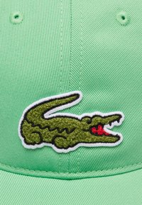 Gorra verde hecha de tela texturizada que presenta un gran logo de cocodrilo verde, delineado en blanco con un acento rojo en su boca.
