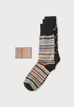 Une paire de chaussettes noires à rayures horizontales multicolores, à côté d’un portefeuille assorti à rayures, sur un fond clair.