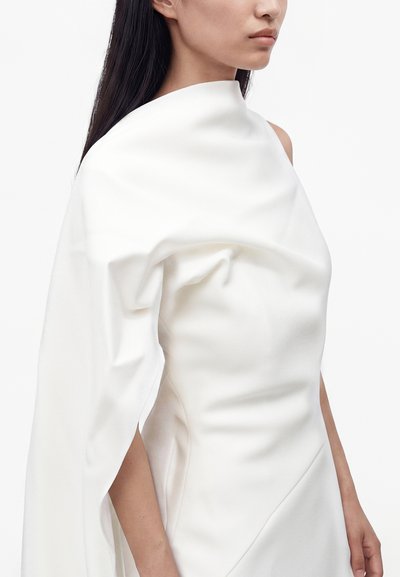 Femme portant une robe blanche à une épaule avec un détail en tissu drapé sur un bras, sur un fond clair uni.