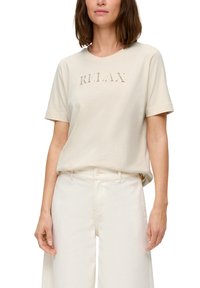 Beige Baumwoll-T-Shirt mit kurzen Ärmeln, verziert mit dem Schriftzug "RELAX" in ausgeschnittenem Lettering. Kombiniert mit hellen, weiten Hosen.