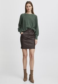 Pull en tricot vert avec texture côtelée, associé à une jupe en cuir marron ajustée. Des bottes chelsea marron complètent la tenue. Design simple, look moderne.