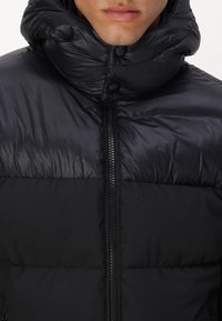 Giacca imbottita nera con colletto alto e chiusura a bottoni. Presenta una zip frontale e una texture liscia e lucida.