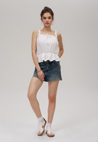 Hvit peplum topp med volanger på skulderstroppene og snor, sammen med blå denimshorts. Modellen har på seg hvite og brune sneakers med ankelstrømper.