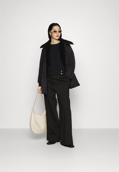 Giubbotto puffy nero con colletto in pelliccia sintetica, maglione lavorato a maglia con dettagli a bottoni, pantaloni neri a gamba larga e borsa a spalla color crema. Occhiali da sole leggeri.