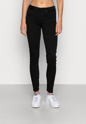 Jeans ajustados negros con una textura suave, dos bolsillos frontales, cierre de botón y detalles de costura. Combinados con zapatillas deportivas blancas.