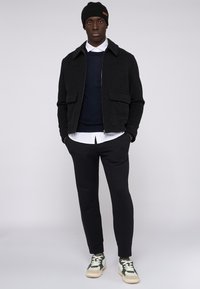 Svart ullblandad jacka med krage, marinblå tröja under, vit skjorta med krage, svarta byxor och sneakers med gröna accenter.