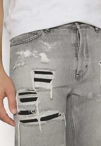 Lähikuva vaaleanharmaista revityistä farkuista, joiden alla on mustat kangaspalat repeämien kohdalla, yhdistettynä valkoiseen yläosaan ja vasemman käden osan näkyessä.