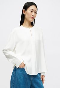 Bílá blůza s dlouhým rukávem a kulatým výstřihem, s malým zlatým akcentem na hrudi. Hladká tkanina, volný střih a boční rozparky.