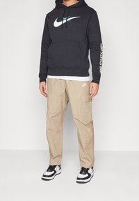 Sweat à capuche gris avec logo Nike blanc, pantalon cargo beige avec logo, et baskets noir et blanc. Design à capuche avec poche avant et manches à motifs.