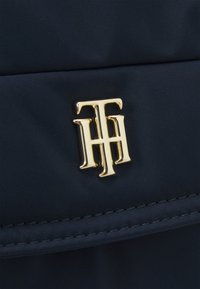Gyllene metallinitialer "TH" logotyp som är fäst vid mörkblå quiltad tyg med synliga sömmar.