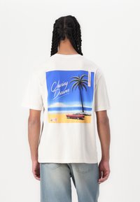 Krem majica s šarenim retro grafičkim dizajnom na leđima. Dizajn uključuje palmu i klasični automobil nasuprot pozadini plaže.
