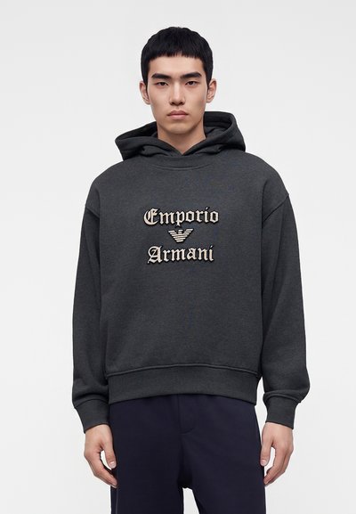 Млад мъж, облечен в тъмносив суитшърт Emporio Armani с голямо лого на гърдите, стоящ пред обикновен светъл фон.