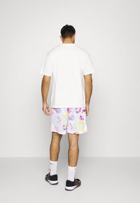 Weiße Baumwoll-T-Shirts kombiniert mit mehrfarbigen Batik-Mesh-Shorts in lila, pink und gelben Farbtönen, getragen mit weißen Socken und schwarzen Schuhen.