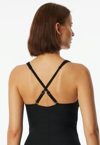 Der schwarze Badeanzug besteht aus einem strukturierten Stoff, hat verstellbare, sich kreuzende Träger und runde goldene Akzente auf der Rückseite. Glatte Silhouette.