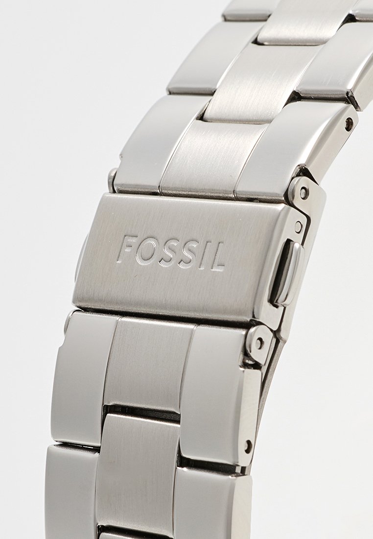 Bracelet de montre en acier inoxydable argenté avec des maillons brossés et polies, arborant un logo Fossil sur le fermoir et une boucle rabattable en métal.