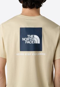 Krémové bavlněné tričko s námořnickým čtvercovým grafikou a logem "THE NORTH FACE" s textem "NEVER STOP EXPLORING" v bílé barvě pod ním. Krátké rukávy.