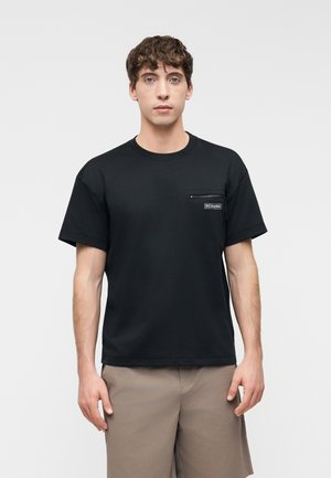 Muškarac u crnoj majici kratkih rukava s džepom na prsima na patentni zatvarač i bež kratkim hlačama, stoji ispred jednobojne svijetle pozadine.