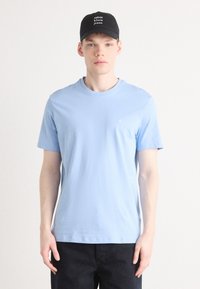 Light blue cotton short-sleeve t-shirt with a round neckline, featuring a small "CK" logo on the chest. Worn with a black cap.