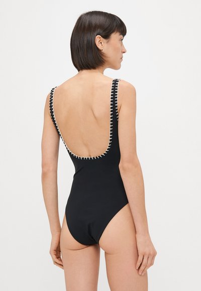 Žena s krátkymi tmavými vlasmi v čiernych jednodielnych plavkách s bielymi švovými detailmi, stojaca chrbtom k fotoaparátu na jemnom svetlom pozadí.