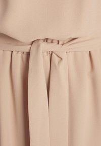 Vestido beige claro con una tela suave y texturizada. Presenta un cinturón atado en la cintura y un detalle plisado debajo del cinturón para añadir dimensión.