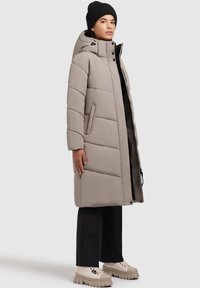 Beige puffer coat with a hood, featuring quilted design, side pockets, and a full zipper. Paired with black pants and chunky footwear.