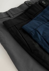 Három összehajtott nadrág szürke, fekete és kék színben, átfedésben elrendezve, látható derékpántokkal, övhurkokkal és gombokkal, egy világos felületen.