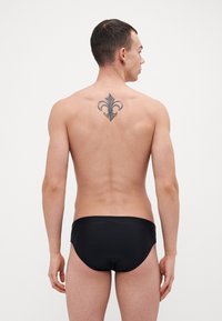 Fato de banho preto com uma textura suave, usado por um modelo masculino de costas, exibindo uma tatuagem floral na parte superior das costas.