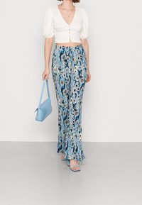 Donna che indossa un crop top bianco e pantaloni larghi blu con fantasia floreale, mentre tiene una borsa blu chiaro e indossa sandali con tacco blu.