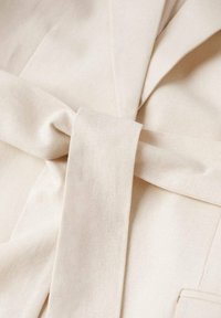 Ceinture en tissu beige nouée en boucle, mettant en avant une texture lisse et des lignes épurées avec un subtil éclat sur un fond de tissu uni.